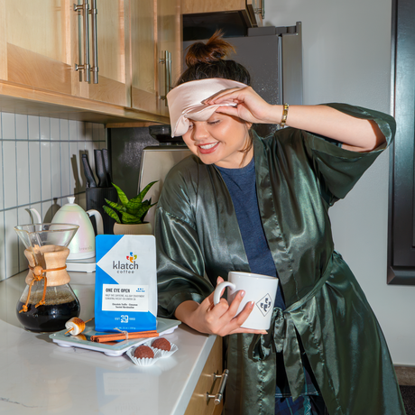 One Eye Open bag on scale next to chemex of coffee on a white counter in a kitchen. Women having her morning coffee with robe and eye mask.