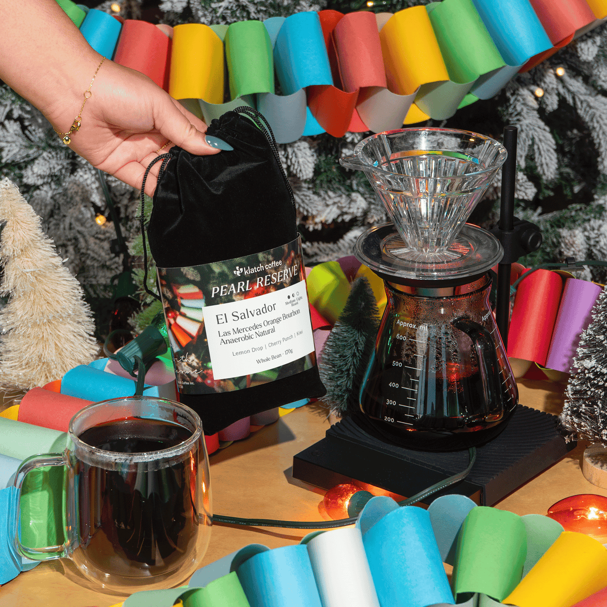 Klatch Coffee Pearl Reserve El Salvador coffee displayed beside a pour-over brewer and glass mug, surrounded by colorful holiday garlands and Christmas decorations.