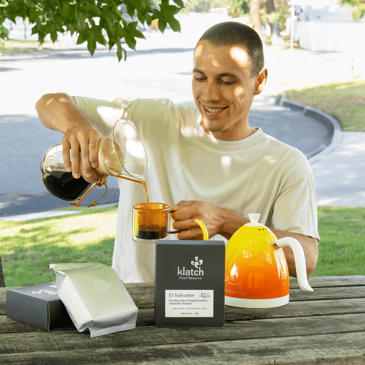 Man pouring freshly brewed coffee from a glass carafe into an amber mug outdoors, with Klatch Pearl Reserve coffee packaging and a bright orange kettle displayed on the table.