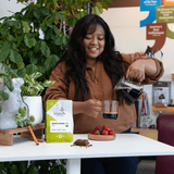 Woman pouring coffee into a cup at a table with a bag of coffee and a coffee tree in the background.