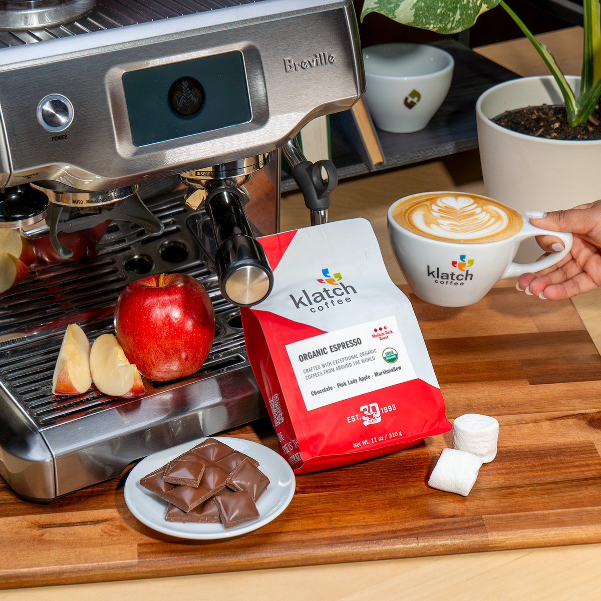 A hand holds a white Klatch Coffee cup with latte art next to a red apple, chocolate pieces, marshmallows, and a bag of Klatch Coffee Organic Espresso on a wooden board, with a Breville espresso machine in the background.
