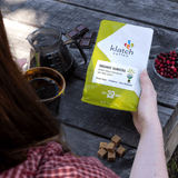 Person holding a Klatch Coffee bag of Organic Sumatra on a wooden table with coffee-making items.