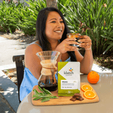 A woman smiling while holding a glass of coffee outdoors at a table, with a bag of Klatch Coffee Organic on a wooden board alongside a pour-over coffee maker, orange slices, chocolate, and green leaves.