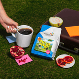 Out of Office Summer blend set on grass with brown brief case, hand holding coffee, guava, pomegranate and slices of blood orange.