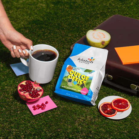 Out of Office Summer blend set on grass with brown brief case, hand holding coffee, guava, pomegranate and slices of blood orange.