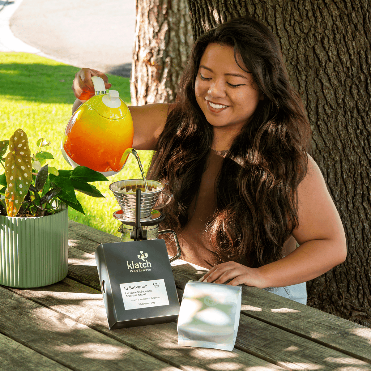 Woman preparing pour over coffee outdoors with a bright orange and yellow kettle, with Klatch Pearl Reserve coffee packaging displayed on a wooden table.