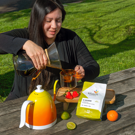 Women pouring coffee with bag of Pacamara Anaerobic Natural on picnic table with strawberries, lime, and orange.