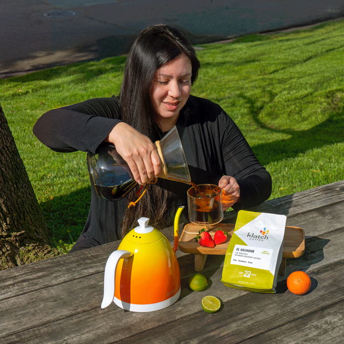 Women pouring coffee with bag of Pacamara Anaerobic Natural on picnic table with strawberries, lime, and orange.