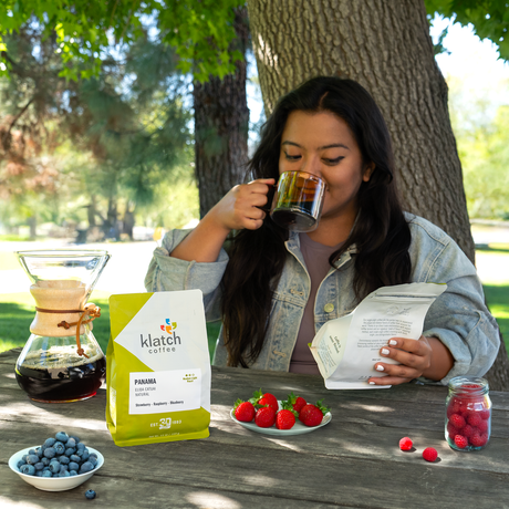 Woman drinking coffee outdoors with a Klatch Coffee Panama Elida Catuai Natuarl bag and berries on a table with chemex pour over.