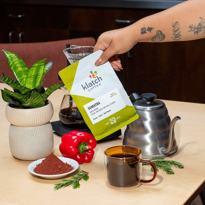 Hand holding a bag of Klatch Coffee Sumatra beside a pour-over setup, coffee mug, red bell pepper, cacao powder, greenery, and a potted plant.