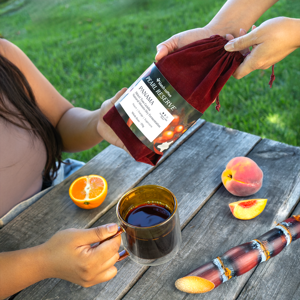Woman handing over a red velvet bag of Altieri Tom Geisha. Other women holding coffee glass mug with fruit on picnic table.