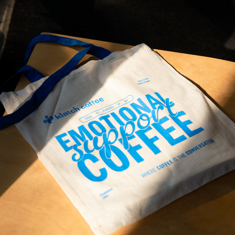 White tote bag with blue text 'Emotional Support Coffee' on a wooden surface.