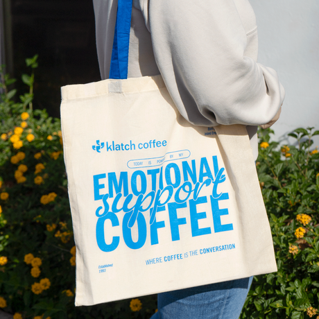 Person holding a tote bag with 'Emotional Support Coffee' text, brand 'klatch coffee' visible, against a blurred natural background.
