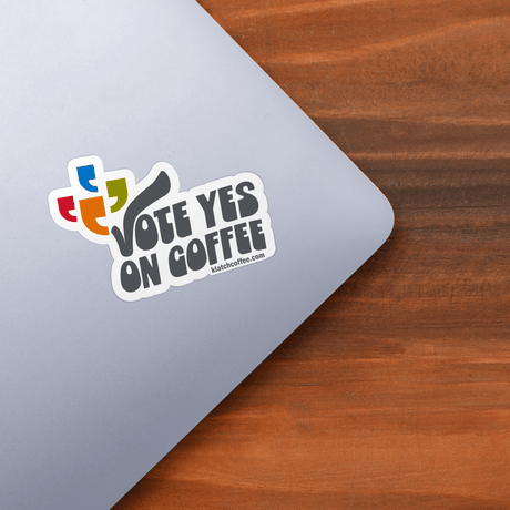 Sticker with 'Vote Yes on Coffee' text on a laptop corner with a wooden surface