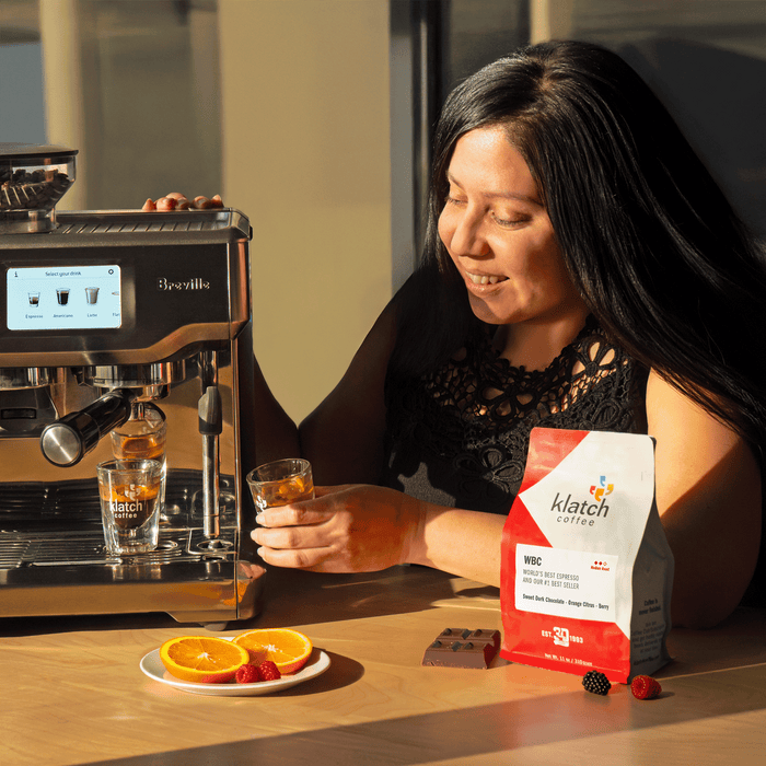 Woman enjoying a espresso drink next to a Breville espresso machine and a bag of klatch coffee WBC, plate of sliced oranges, raspberries, and a chocolate bar on table.
