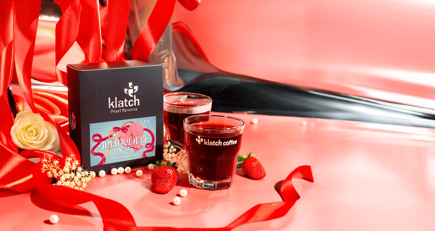 Klatch Coffee Pearl Aphrodite box displayed on red ribbons, surrounded by roses, elderflowers, and strawberries on a pink background.
