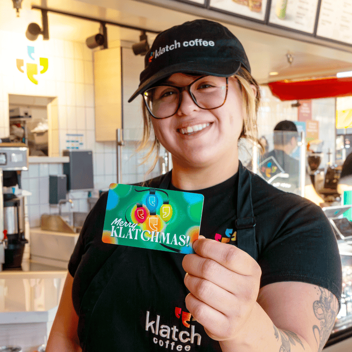 A barista stands in a café holding a Christmas gift card printed with the message “Merry Klatchmas.”