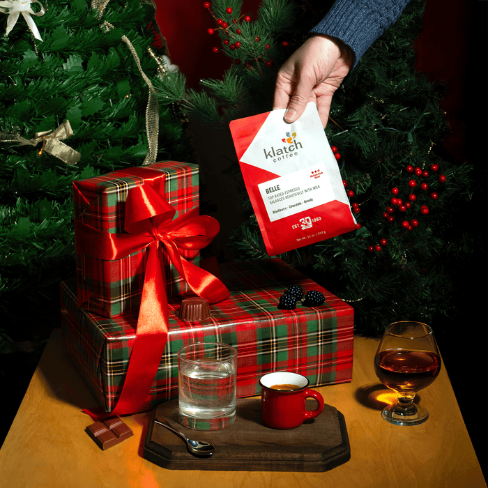 Person holding a Klatch coffee Belle Espresso with Christmas presents and drinks on a table.