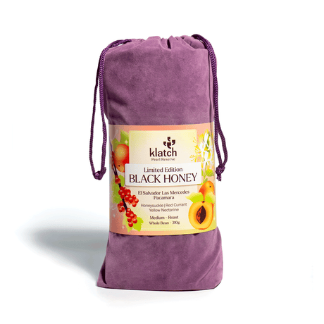 Purple velvet drawstring bag with a wraparound label reading “Limited Edition Black Honey El Salvador Las Mercedes Pacamara,” decorated with illustrations of honeysuckle, red currants, and yellow nectarines on a white background.