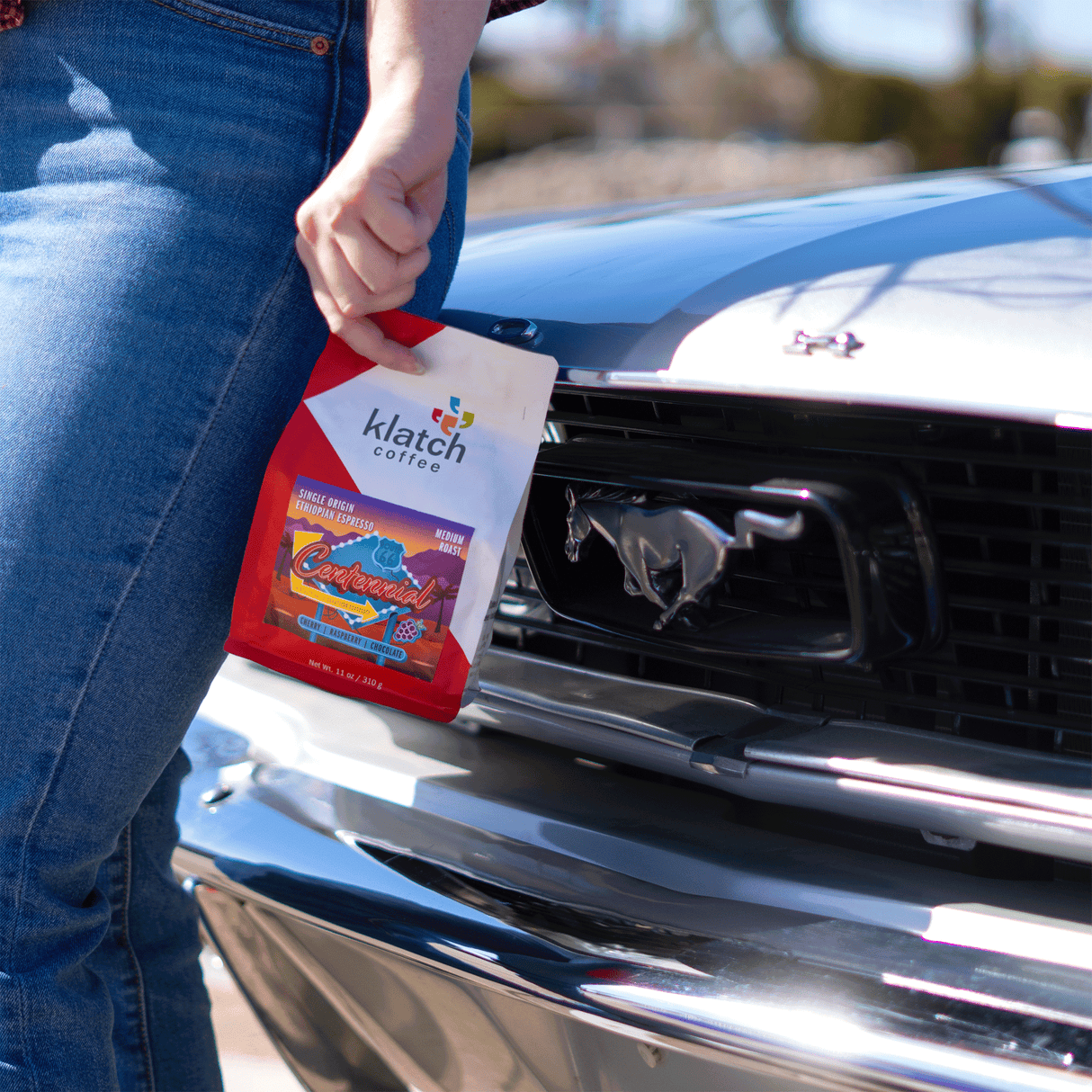 Person holding a Klatch Coffee Centennial bag in front of a Ford Mustang.