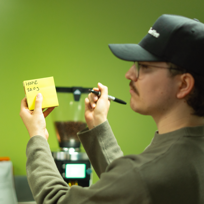Man holding a yellow notepad with 'house 20g' written on it, standing in front of a green wall.