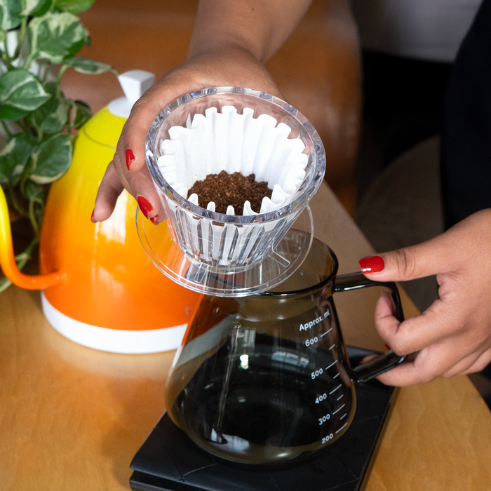 A person holds a TIMEMORE Crystal Eye B75 Dripper above a coffee server, with a plant and and orange kettle blurred in the background.