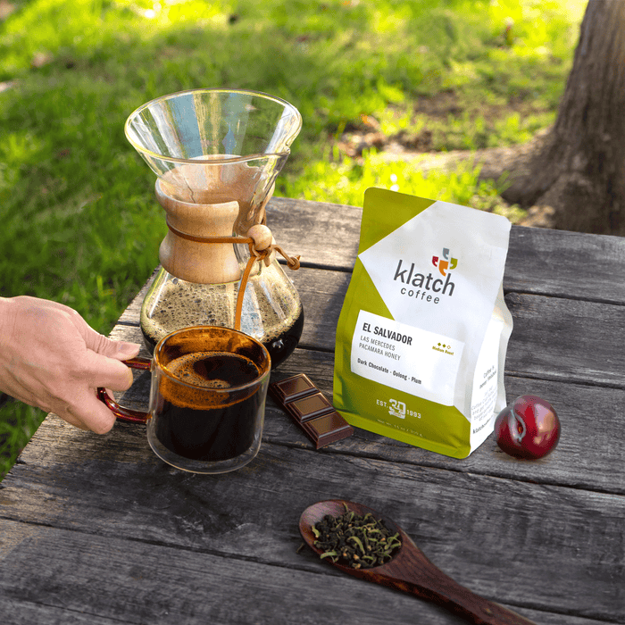 Person pouring coffee from a Chemex into a glass mug on a wooden table with a Klatch Coffee bag, plum, dark chocolate bar an loose oolong tea on table.