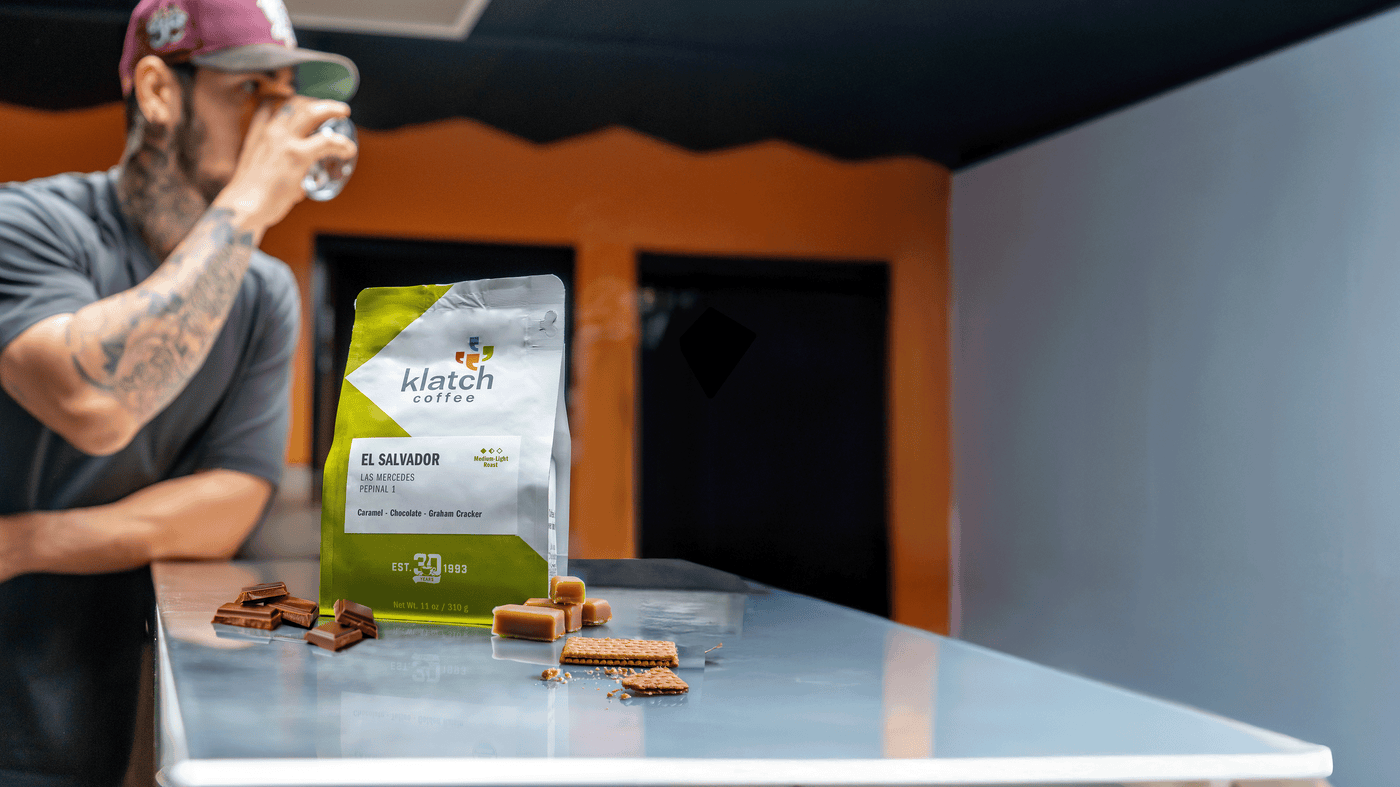 Man drinking from a glass with a Klatch Coffee El Salvador Las Mercedes Pepinal 1 bag with caramel candies, chocolate stacked and graham cracker on a table.