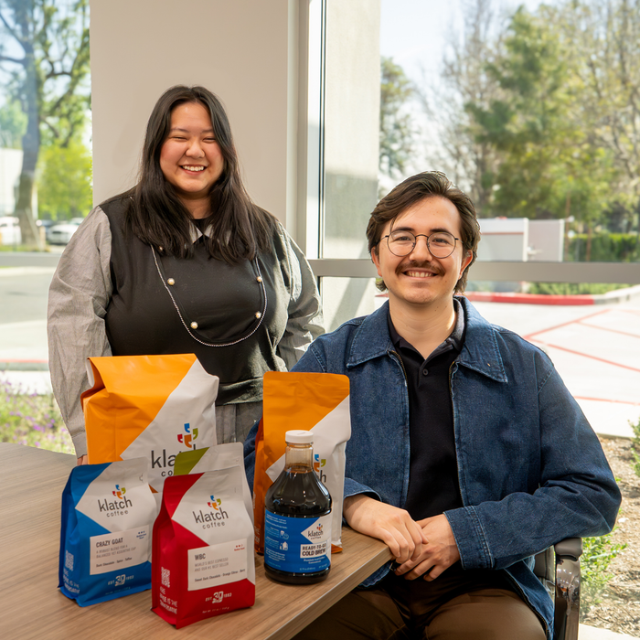 Man and woman smiling at the camera in front of a window, with coffee bags displayed on a table in front of them.