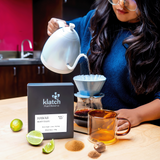 Person pouring tea from a white kettle, with tea bag and limes on a wooden table.