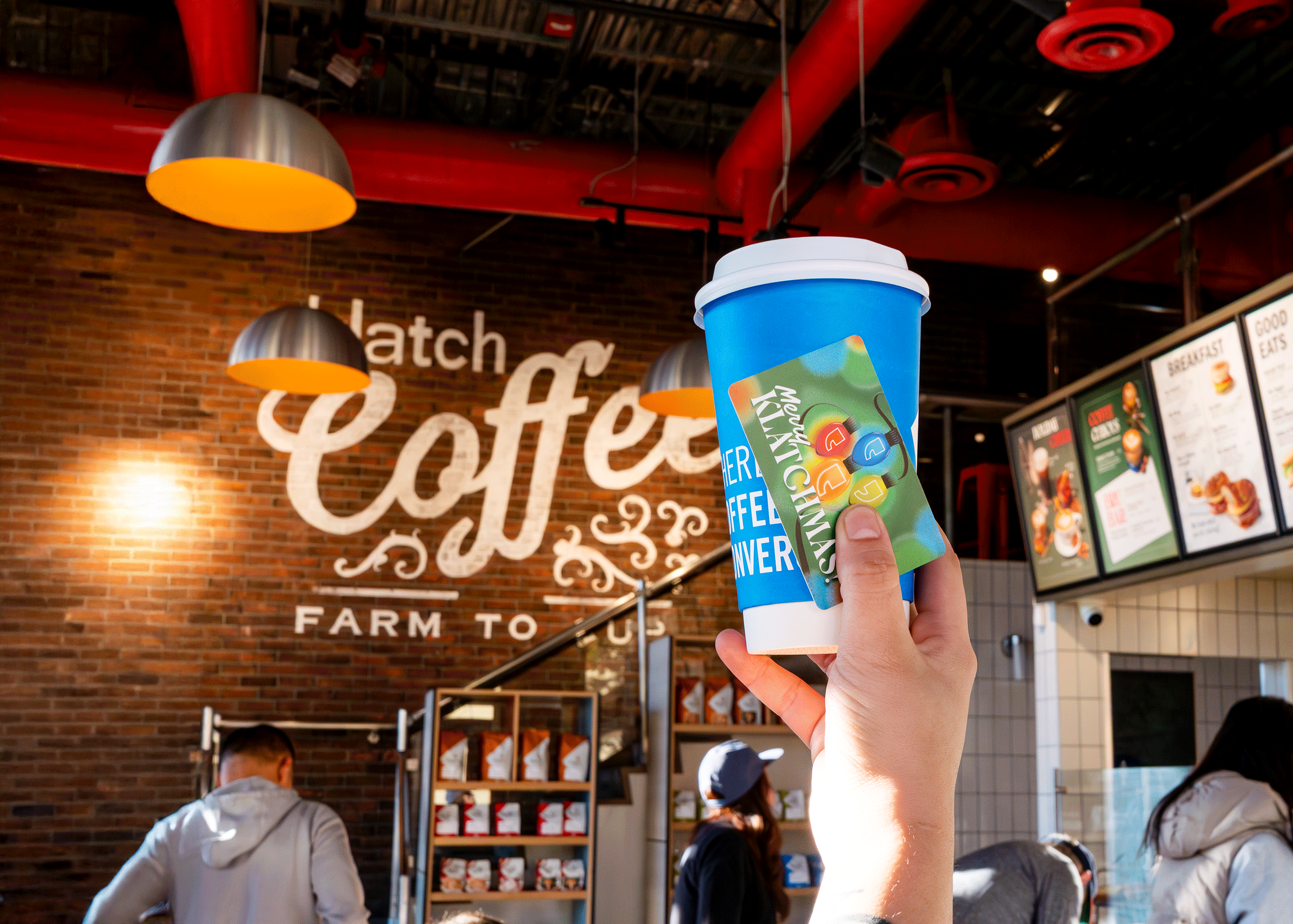 Person holding a blue coffee cup and a holiday gift card that says “Merry Klatchmas” in a café setting.