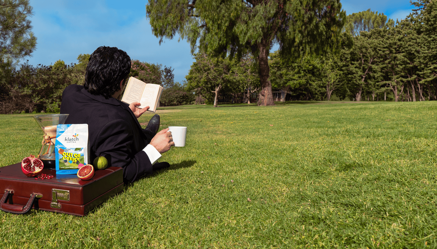 Person sitting on grass reading a book with a briefcase, a chemex of coffee on top with a bag of Out of Office and fruit.
