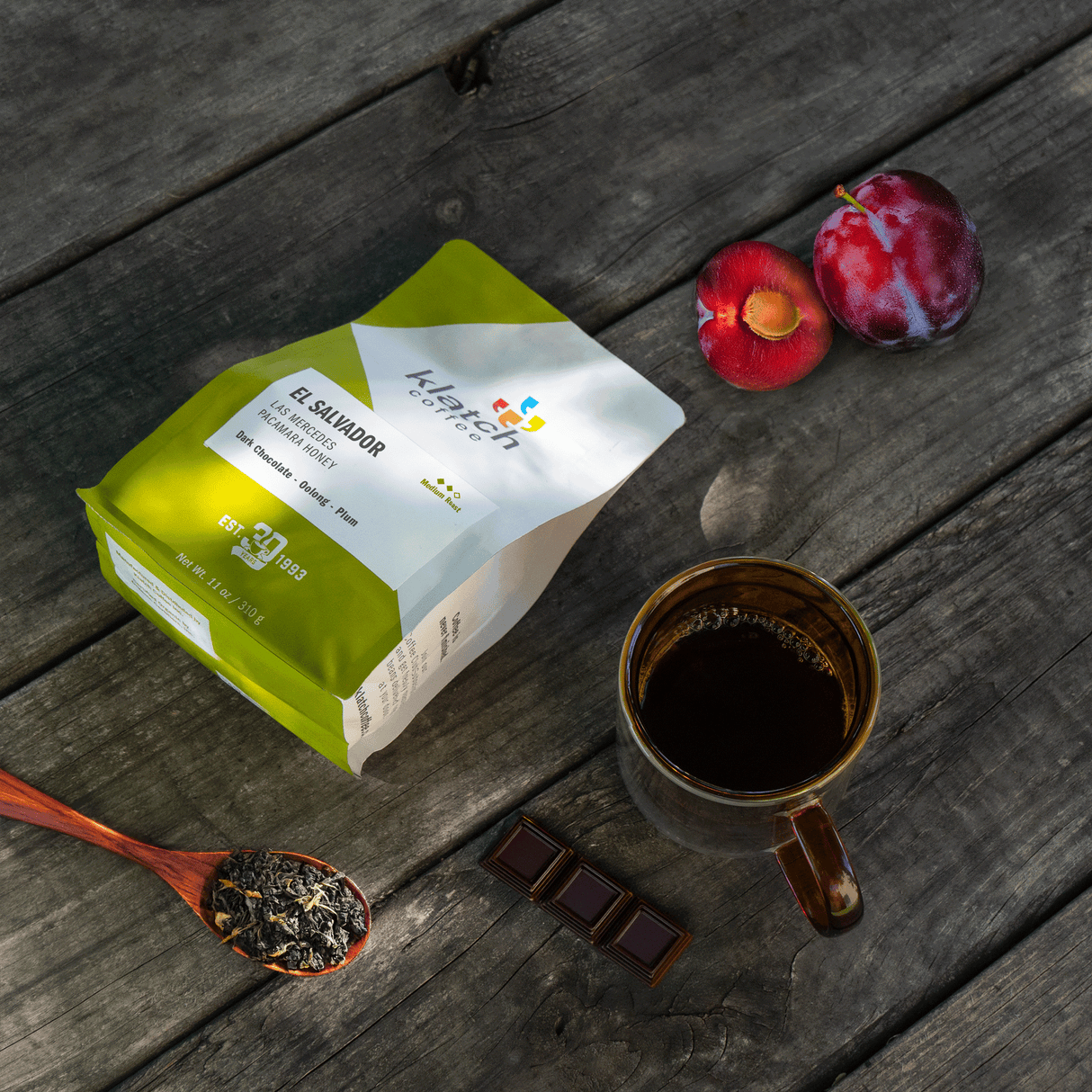 Klatch coffee package with a cup of coffee, dark chocolate bar, plums, and a wooden spoon with dry oolong on a wooden picnic table.