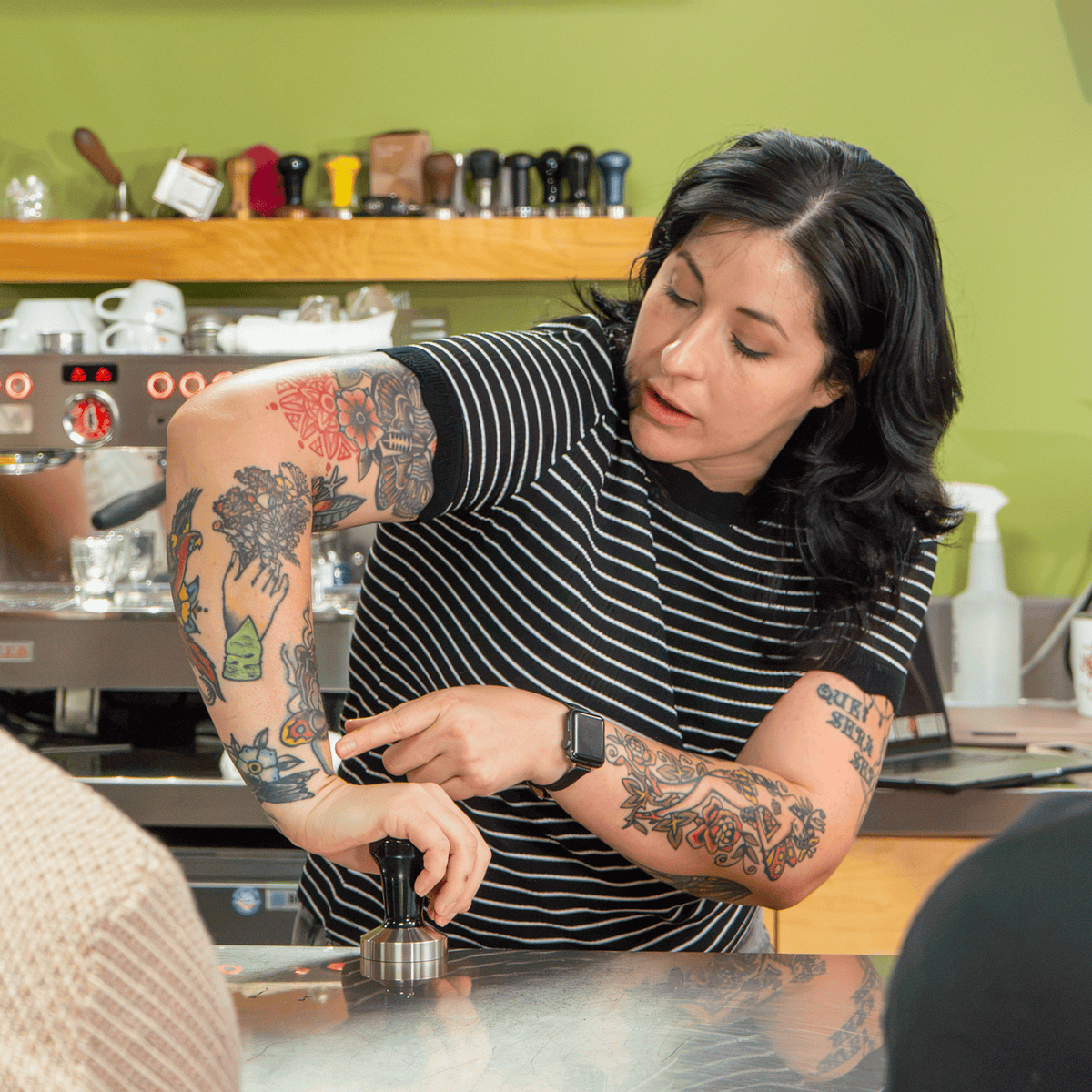 Barista demonstrating espresso preparation by tamping coffee at a café counter, with an espresso machine in the background.
