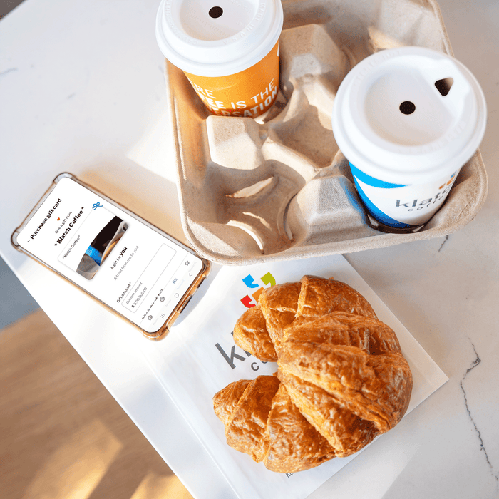 A smartphone showing the Klatch Coffee mobile gift card app, placed on a table beside a croissant and two cups of coffee.