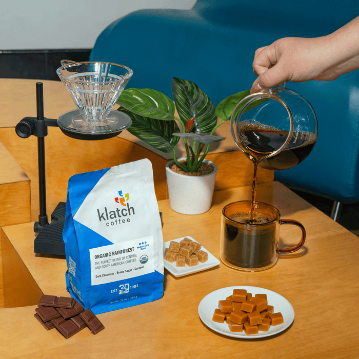 Klatch Coffee Organic Rainforest blend beside a pour-over setup as freshly brewed coffee is poured into a mug, surrounded by dark chocolate, brown sugar, caramel, and a potted plant.