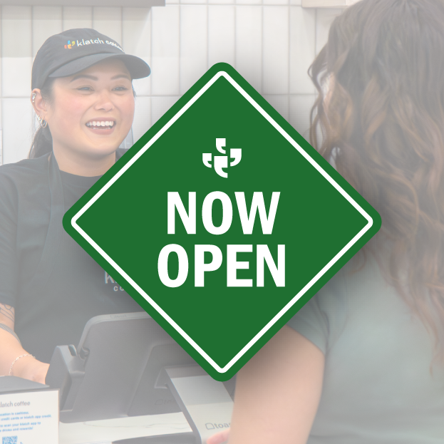 Klatch Coffee barista smiling with a customer behind a green “Now Open” graphic.
