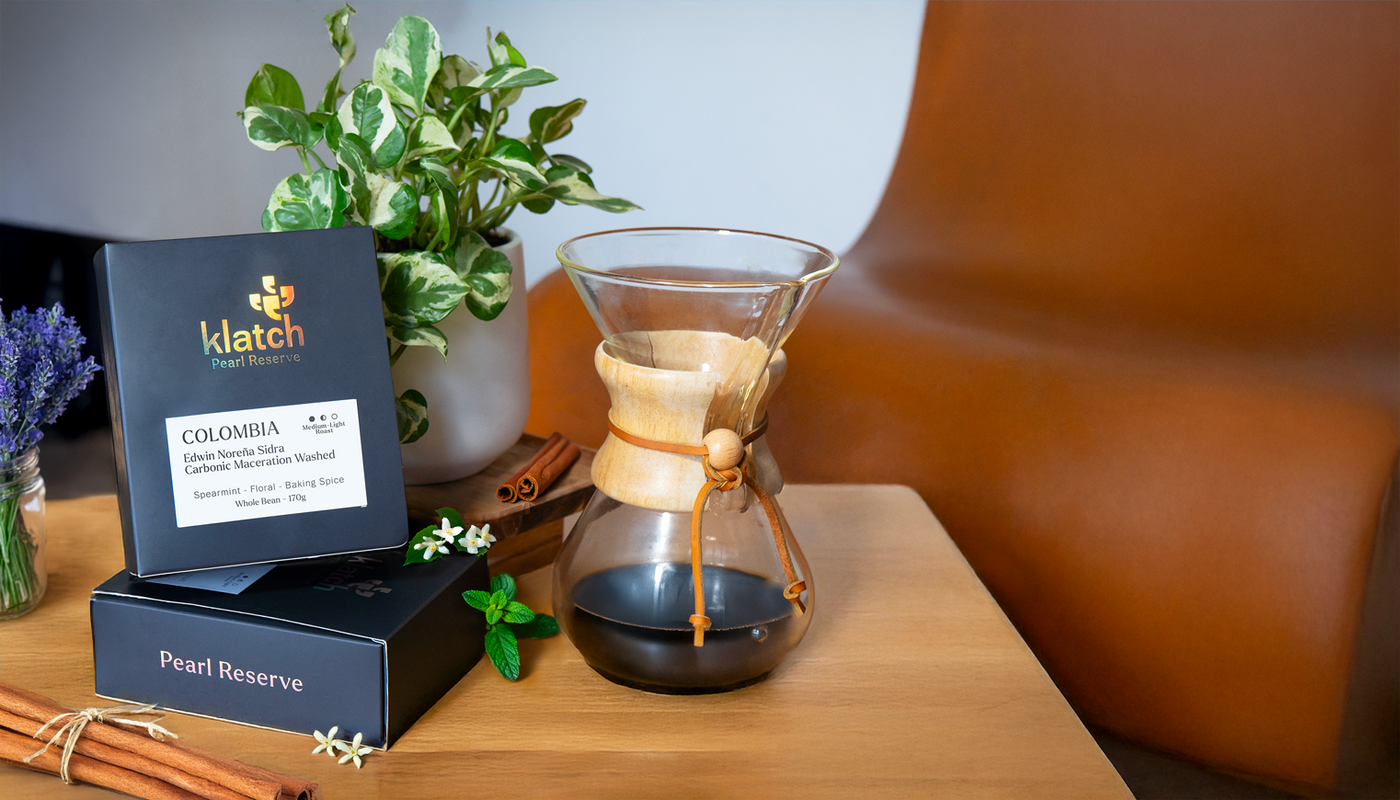 Grey pearl box stacked on another by a plant on a wooden stool, next to a Chemex of coffee with cinnamon sticks and spearmint, in front of a brown chair.