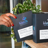 Person holding a Klatch Pearl Reserve coffee box  of Colombia Edwin Norena Sidra Carbonic Maceration Washed on a shelf with a plant in the background.