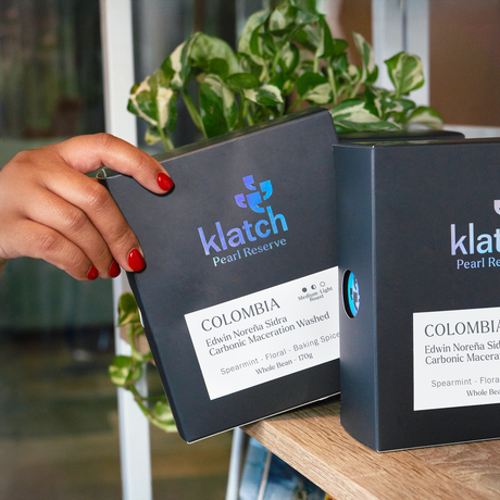 Person holding a Klatch Pearl Reserve coffee box  of Colombia Edwin Norena Sidra Carbonic Maceration Washed on a shelf with a plant in the background.