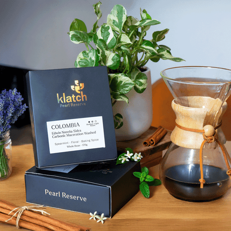 Klatch Coffee Pearl box stacked on another by a plant on a wooden stool, next to a Chemex of coffee with jar of lavender, cinnamon sticks, jasmine flowers and spearmint, in front of a brown chair.