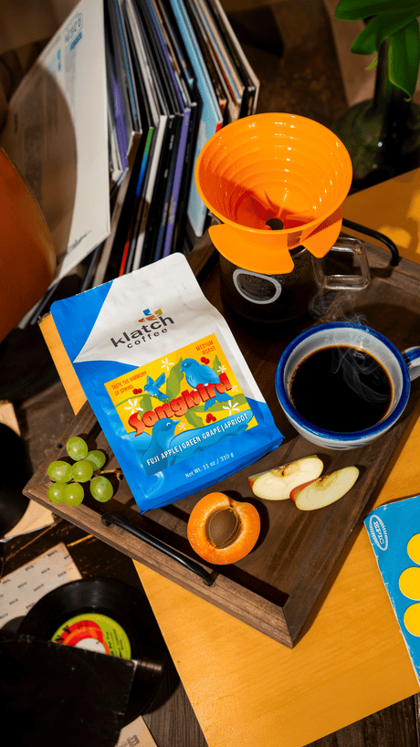 Tray with Songbird bag of coffee, with grapes, apricot and slices of apple on a table surrounded records open and closed.