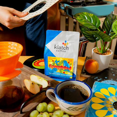 Person holding 45 record with a table in front of him. A tray of Songbird  with a pour over, a bag of coffee, grapes, apricot and slices of apple on a table surrounded records, record player in background.