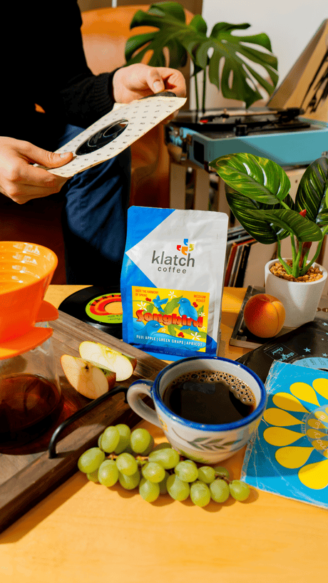 Person holding 45 record with a table in front of him. A tray of Songbird  with a pour over, a bag of coffee, grapes, apricot and slices of apple on a table surrounded records, record player in background.