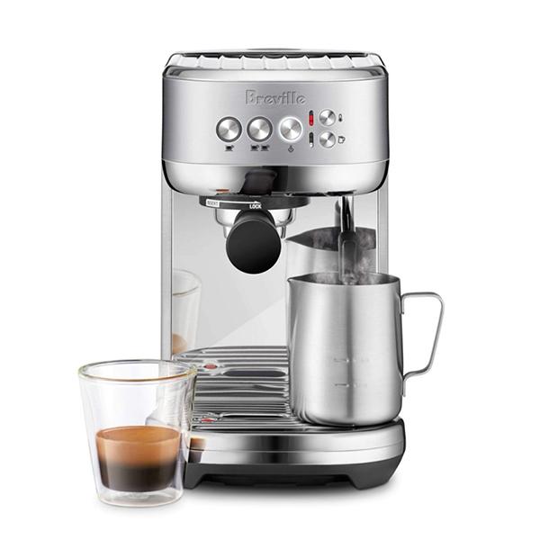 Breville Bambino Plus espresso machine with portafilter, steam wand, stainless steel milk pitcher, and tamper on a white background next to a cup of espresso. 